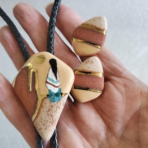 May include: A bolo tie and matching earrings set. The bolo tie features a diamond-shaped pendant with a Native American figure design, accented with gold-colored details. The earrings have a similar design. The set is made of ceramic and has a leather cord.