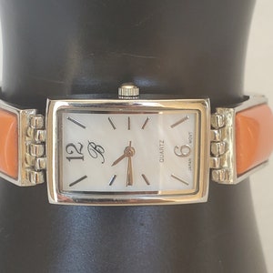 May include: A silver watch with a white face and orange band. The watch face has black Roman numeral markings and the text "QUARTZ" and "JAPAN MOV'T". The watch face also has a small "B" logo.