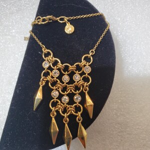 May include: A gold-tone chain necklace featuring a multi-layered link design adorned with clear round crystals. The pendant section includes five dangling, smooth, diamond-shaped gold-tone charms. A small circular tag is attached to the chain.