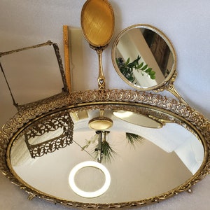 May include: A gold-toned vanity tray with an oval mirror and ornate detailing. The tray has two smaller mirrors, one oval and one round, and a comb.