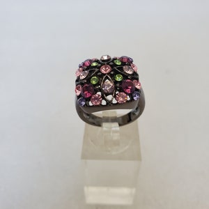 May include: A black ring with a square-shaped setting featuring a cluster of pink, purple, and green rhinestones.