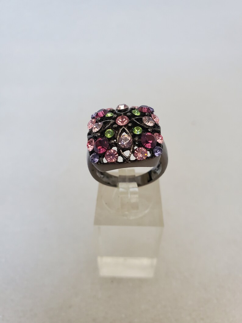 May include: A black ring with a square-shaped setting featuring a cluster of pink, purple, and green rhinestones.