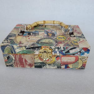 May include: A rectangular box with a bamboo handle and a gold-colored clasp. The box is covered in a collage of vintage travel labels and stamps in various colors. The handle is made of curved bamboo.