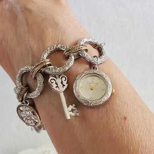 May include: A silver-toned charm bracelet with a watch, key, and decorative charms. The watch has a gold-tone bezel and a cream-colored face. The bracelet links are adorned with clear crystals and gold-tone accents.