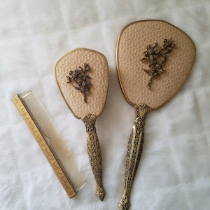 May include: A vintage vanity set featuring a hand mirror, a smaller mirror, and a comb. The set has a gold-tone finish with floral accents and a woven fabric backing. The handles and comb are intricately detailed.