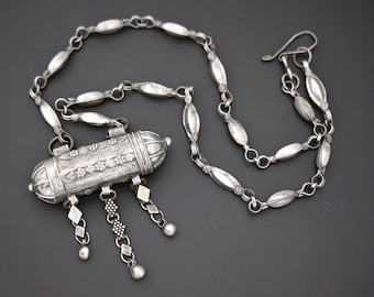 May include: A silver necklace with a cylindrical pendant and decorative chain links. The pendant is adorned with floral and geometric patterns, and dangling charms. The necklace is displayed on a dark gray background.
