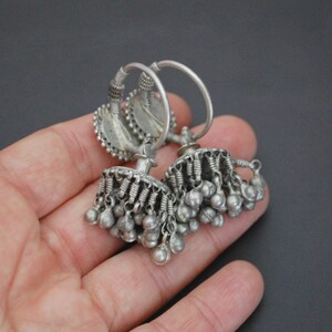 May include: A pair of silver hoop earrings with dangling charms. The earrings have a circular design with a small, round charm in the centre. The charms are made of silver and have a bell-like shape.