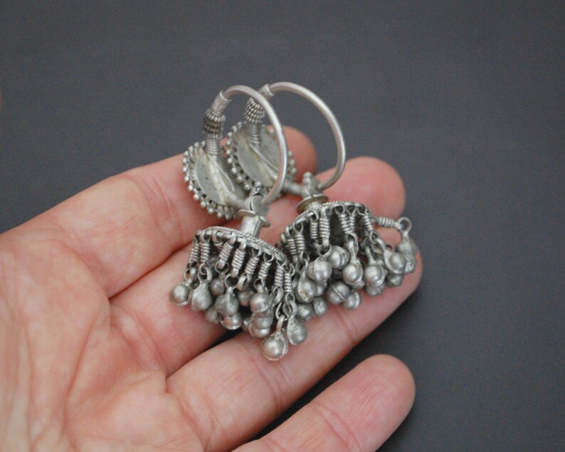 May include: A pair of silver hoop earrings with dangling charms. The earrings have a circular design with a small, round charm in the centre. The charms are made of silver and have a bell-like shape.