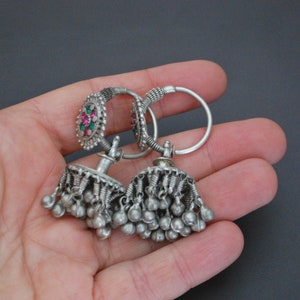 May include: A pair of silver earrings with dangling charms. The earrings feature a small round design with pink and green stones.