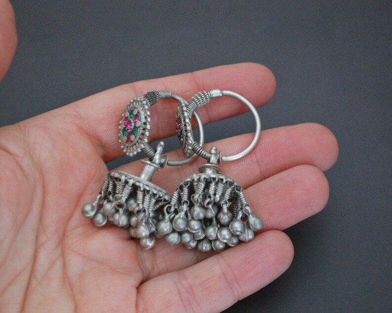 May include: A pair of silver earrings with dangling charms. The earrings feature a small round design with pink and green stones.