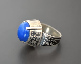 Può includere: Un anello d'argento con una pietra preziosa blu incastonata in una montatura a castone. La fascia è decorata con un intricato motivo floreale.
