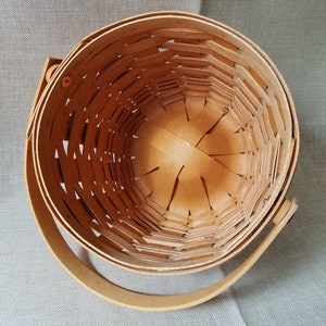 Puede incluir: Una cesta de mimbre de madera con un acabado natural y un solo asa. La cesta est&aacute; vac&iacute;a y tiene forma redonda.