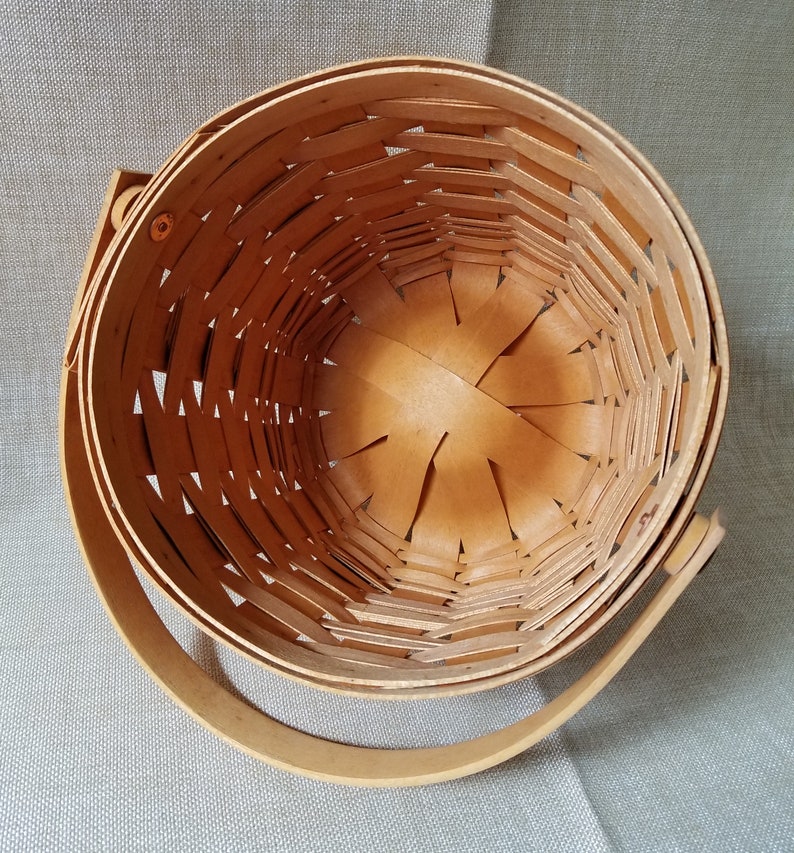 Puede incluir: Una cesta de mimbre de madera con un acabado natural y un solo asa. La cesta est&aacute; vac&iacute;a y tiene forma redonda.