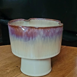 May include: A ceramic planter with a wide, rounded top and a pedestal base. The planter is glazed in shades of brown, purple, and cream, creating a gradient effect.
