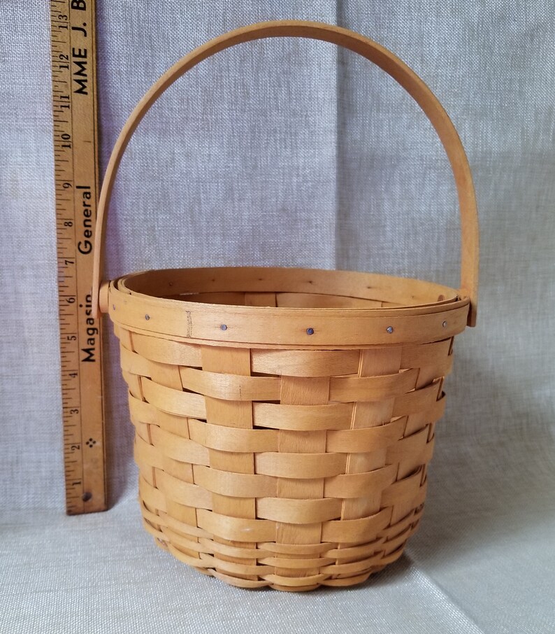Puede incluir: Una peque&ntilde;a cesta tejida natural con un solo asa. La cesta est&aacute; hecha de madera marr&oacute;n claro y tiene un patr&oacute;n tejido. La cesta mide aproximadamente 15 cm de alto.