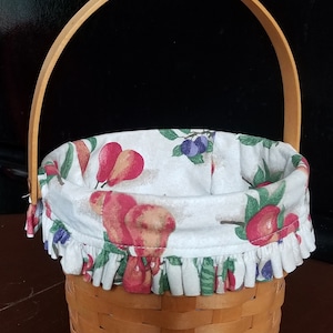 May include: A brown wicker basket with a fabric liner featuring a fruit print. The liner has a ruffled edge and is white with red, green, and blue fruit designs. The basket has a wooden handle.