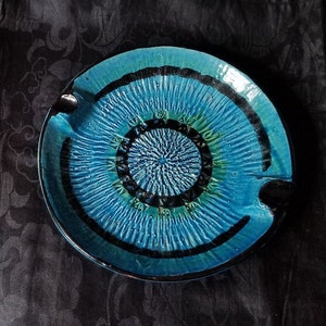 May include: A blue and black ceramic ashtray with a decorative design. The ashtray is circular and has a raised rim.