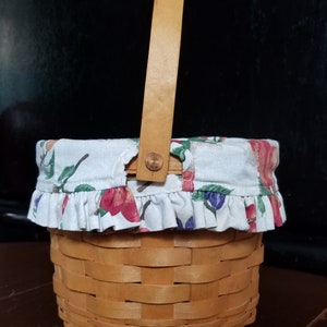 Puede incluir: Una peque&ntilde;a cesta de madera con un forro de tela. El forro es blanco con un patr&oacute;n floral de color rosa, rojo, verde y azul. La cesta tiene un asa de madera.