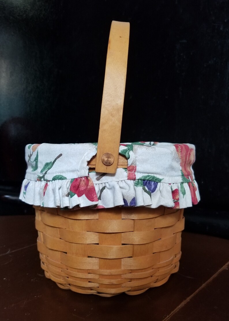 Puede incluir: Una peque&ntilde;a cesta de madera con un forro de tela. El forro es blanco con un patr&oacute;n floral de color rosa, rojo, verde y azul. La cesta tiene un asa de madera.