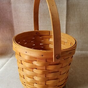 Puede incluir: Una cesta peque&ntilde;a, redonda y tejida con un acabado de madera natural y un solo asa. La cesta est&aacute; vac&iacute;a.