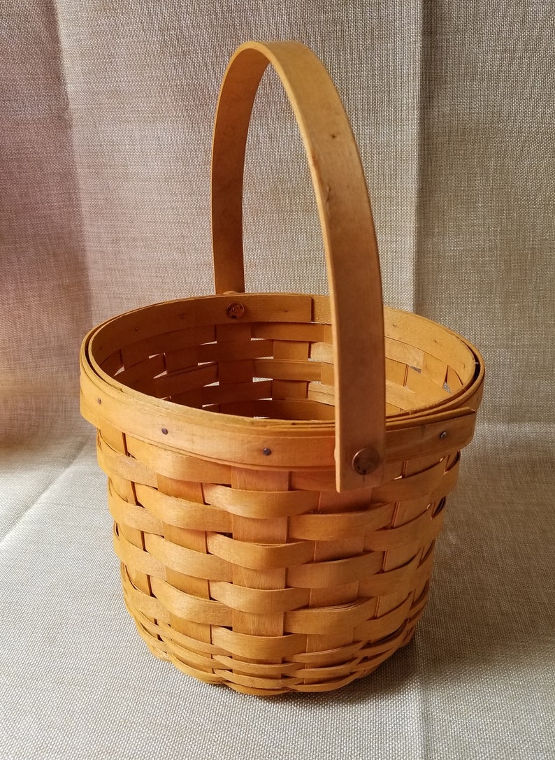Puede incluir: Una cesta peque&ntilde;a, redonda y tejida con un acabado de madera natural y un solo asa. La cesta est&aacute; vac&iacute;a.