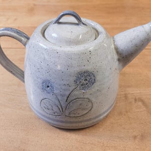 Peut inclure: Une théière en céramique blanche avec un motif floral bleu. La théière a un corps rond et un bec verseur sur le côté. Le couvercle a un bouton sur le dessus.