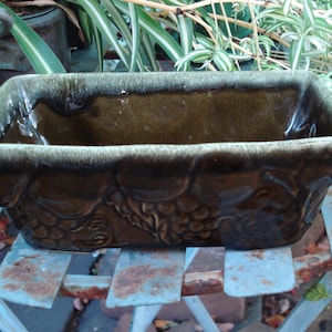 May include: A rectangular green glazed ceramic planter with a grapevine design. The planter has a light green glaze on the top edge and a darker green glaze on the bottom.