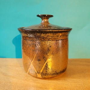 May include: A brown ceramic jar with a lid, featuring a textured, speckled surface and a geometric pattern of gold lines. The jar has a rounded body and a slightly flared opening.