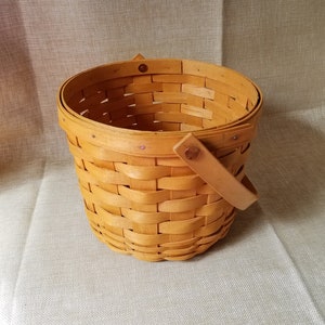 Puede incluir: Una cesta tejida con un solo asa. La cesta est&aacute; hecha de madera natural y tiene un color marr&oacute;n claro.