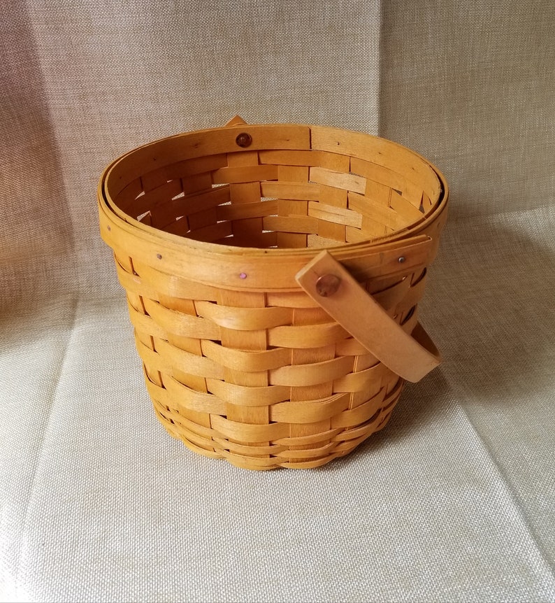 Puede incluir: Una cesta tejida con un solo asa. La cesta est&aacute; hecha de madera natural y tiene un color marr&oacute;n claro.