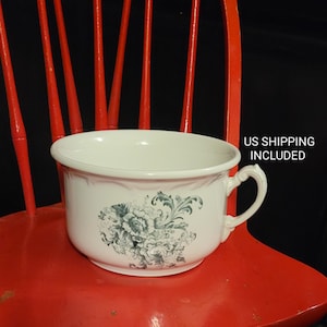 May include: White ceramic chamber pot with a green floral design. The pot has a handle on one side and sits on a red wooden chair.