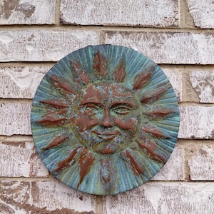May include: A circular wall plaque featuring a relief sculpture of a sun face with radiating rays. The surface has a weathered, oxidized finish with a mix of deep copper-brown tones and teal-green patina, creating a textured, aged appearance.