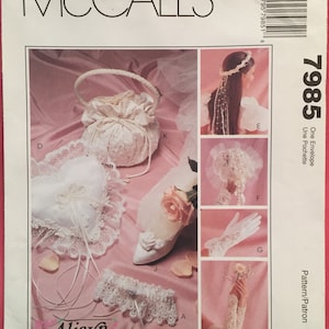 May include: A set of white lace bridal accessories, including a garter, a pillow, a small bag, and a pair of gloves. The accessories are arranged on a pink background. The pattern number 7985 is visible in the upper right corner of the image. The text "One Envelope Une Pochette" is also visible in the upper right corner of the image.