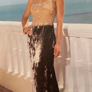 May include: A formal evening gown with a beaded bodice and a dark velvet skirt. The dress has spaghetti straps and a floor-length hemline. The model is standing on a balcony overlooking the ocean. The letter "B" is visible in the image.