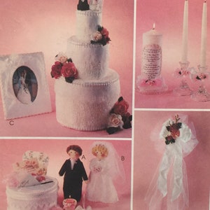 May include: A white wedding cake with a bride and groom topper, a white picture frame with a wedding photo, a white box with a floral design, a white box with a floral design, a white candle with a wedding poem, two white candles, and a white tulle bow with red roses.