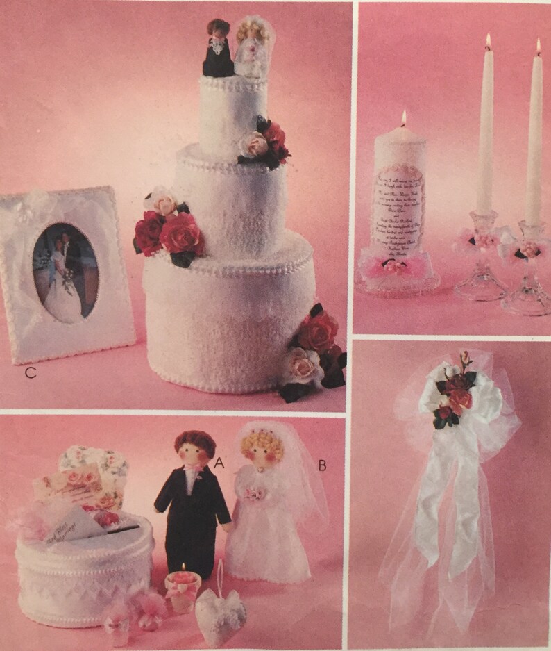 May include: A white wedding cake with a bride and groom topper, a white picture frame with a wedding photo, a white box with a floral design, a white box with a floral design, a white candle with a wedding poem, two white candles, and a white tulle bow with red roses.
