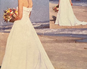 May include: A strapless white wedding dress with a long train. The dress features a fitted bodice and a flowing skirt. The bride is holding a bouquet of colorful flowers. The background shows a beach scene.