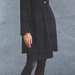 May include: A woman wearing a black coat dress with gold buttons, black tights, and black heels. She is also wearing a gold bracelet. The background is a solid gray color.