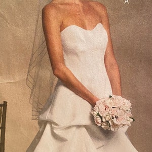 May include: A strapless white wedding dress with a sweetheart neckline and a layered skirt. The bride holds a bouquet of pink flowers. She wears a tiara and earrings. A sheer veil is draped over her hair.