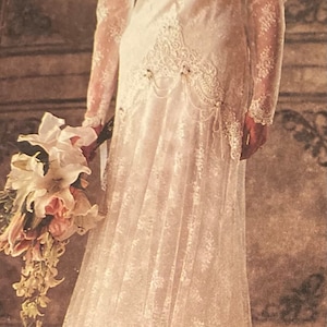 May include: A white floor-length dress featuring long lace sleeves, a V-neckline with intricate lace detailing, and a layered skirt with a scalloped hem. The garment includes a decorative waist panel with hanging beaded accents and a floral hair accessory.