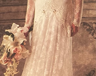 May include: A white floor-length dress featuring long lace sleeves, a V-neckline with intricate lace detailing, and a layered skirt with a scalloped hem. The garment includes a decorative waist panel with hanging beaded accents and a floral hair accessory.