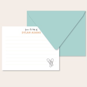 May include: A white lined notepad with a light blue envelope. The notepad has the text "from the desk of DYLAN ADAMS" printed on the top. A simple drawing of two paperclips forming a heart is at the bottom of the notepad.