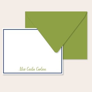 May include: A white card with a navy blue border and the text "Miss Cecilia Carlson" in cursive script. The card is paired with a green envelope.
