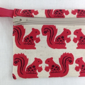 May include: A small zippered pouch with a red fabric exterior featuring a repeating pattern of red squirrels holding acorns. The pouch has a red fabric loop handle.