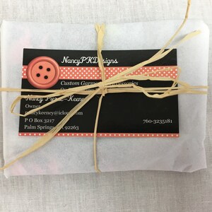 May include: A white paper package tied with a brown raffia ribbon. The package has a black and white business card with the text "Nancy PKD Designs", "Custom Garments & Accessories", "Nancy Fier-Keeney", "Owner", "Pacykeeney@icloud.com", "PO Box 3217", "Palm Springs 92263", and "760-3235181". The business card has a red button with four black holes on it.