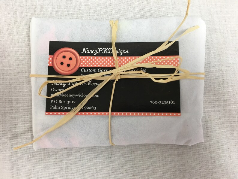 May include: A white paper package tied with a brown raffia ribbon. The package has a black and white business card with the text "Nancy PKD Designs", "Custom Garments & Accessories", "Nancy Fier-Keeney", "Owner", "Pacykeeney@icloud.com", "PO Box 3217", "Palm Springs 92263", and "760-3235181". The business card has a red button with four black holes on it.