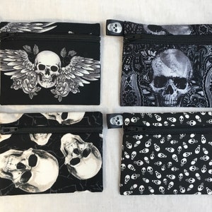 May include: Four small, black zippered pouches with skull designs. One features a skull with wings and roses, another a skull against a paisley background, a third with multiple skulls, and the last with a pattern of small skulls.