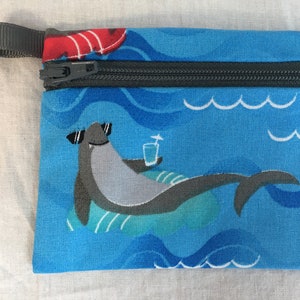 Puede incluir: Una bolsa azul con cremallera con un tiburón que lleva gafas de sol y una bebida, flotando sobre un patrón de olas azules y verdes.