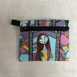May include: A blue and purple zippered pouch with a colorful stained glass pattern featuring a character from the Nightmare Before Christmas. The character has red hair, blue skin, and is wearing a yellow and blue dress.