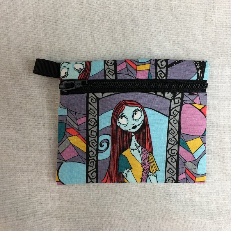 May include: A blue and purple zippered pouch with a colorful stained glass pattern featuring a character from the Nightmare Before Christmas. The character has red hair, blue skin, and is wearing a yellow and blue dress.